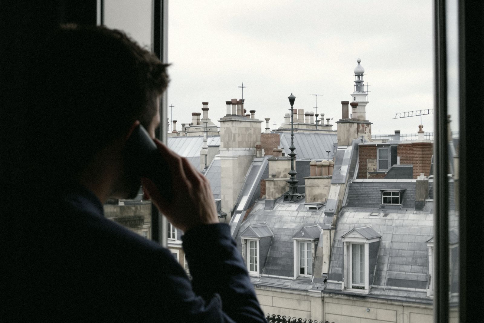 Homme de dos au téléphone devant une fenêtre ouverte sur les toits en zinc de Paris