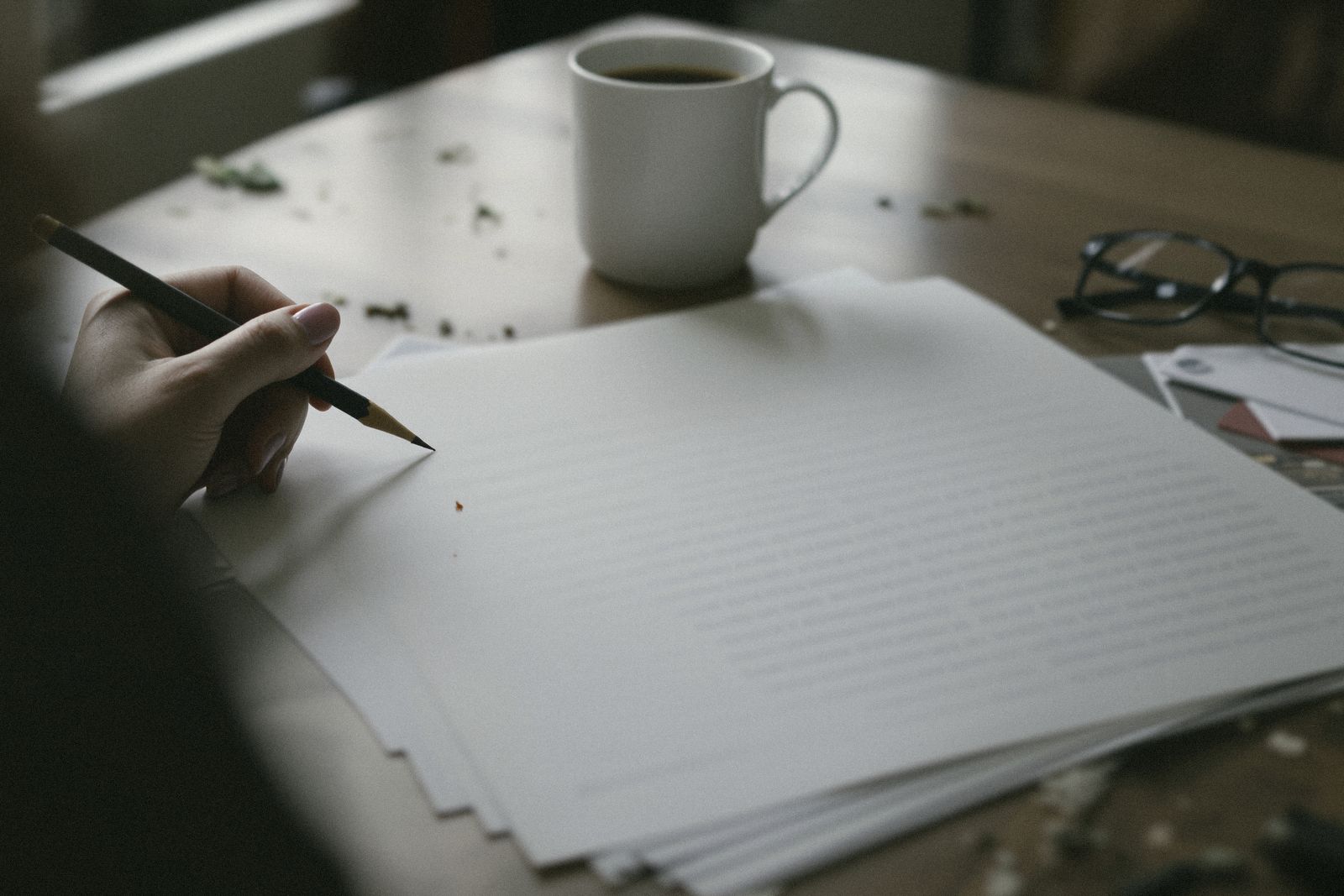 Main qui annote au crayon des feuilles imprimées sur une table en bois, à côté d'une tasse de café