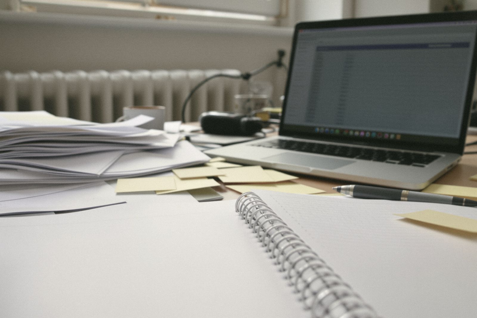 Bureau encombré avec des relevés imprimés, un carnet à spirale, des post-it et un ordinateur portable devant un radiateur