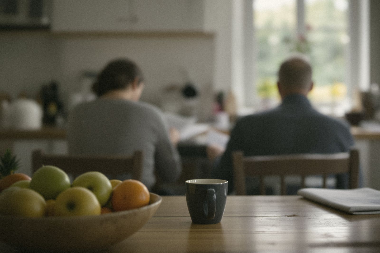Deux personnes de dos, floues, qui consultent des papiers à la table de la cuisine, corbeille de fruits au premier plan