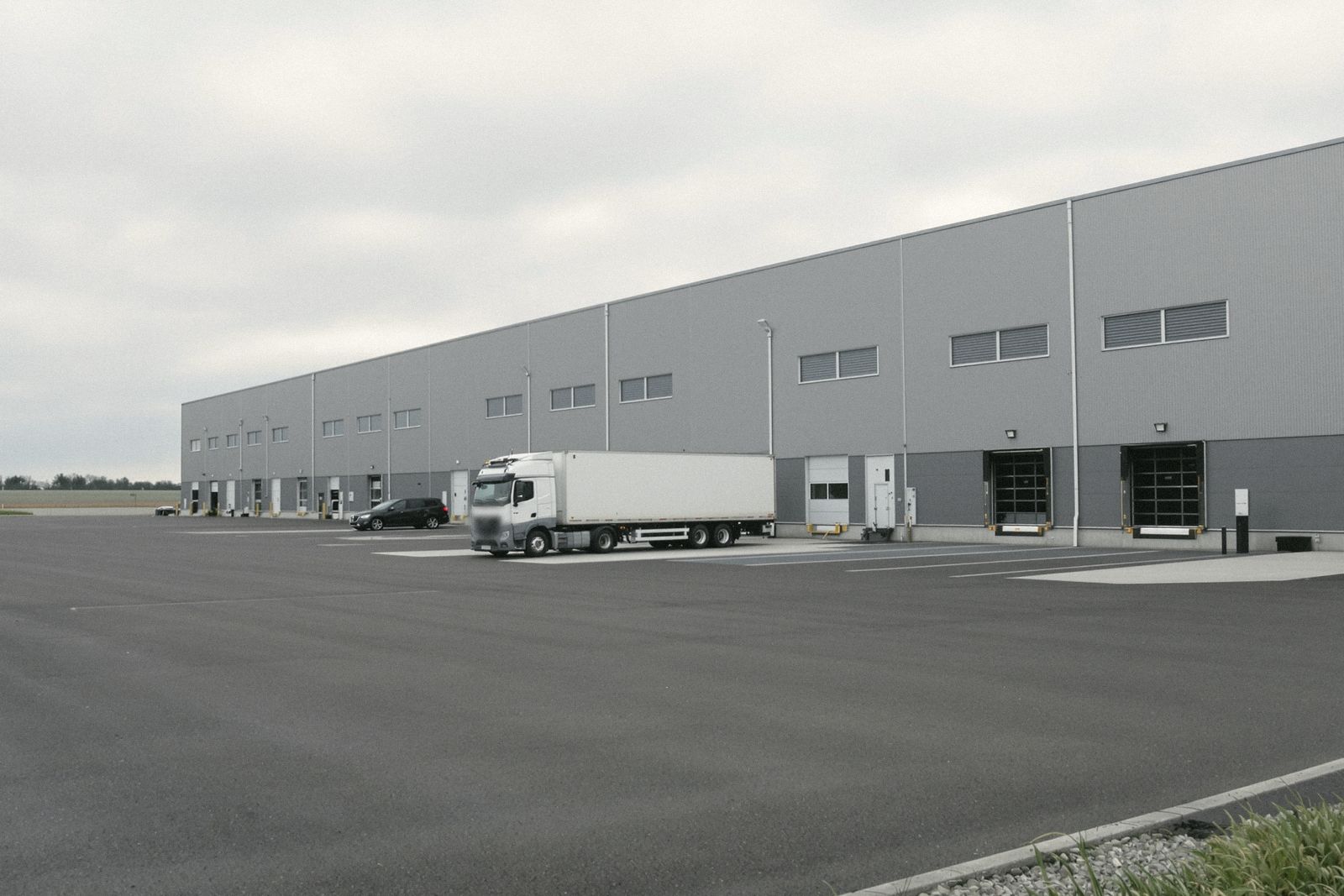 Entrepôt logistique gris avec quais de chargement et un camion blanc stationné, ciel couvert